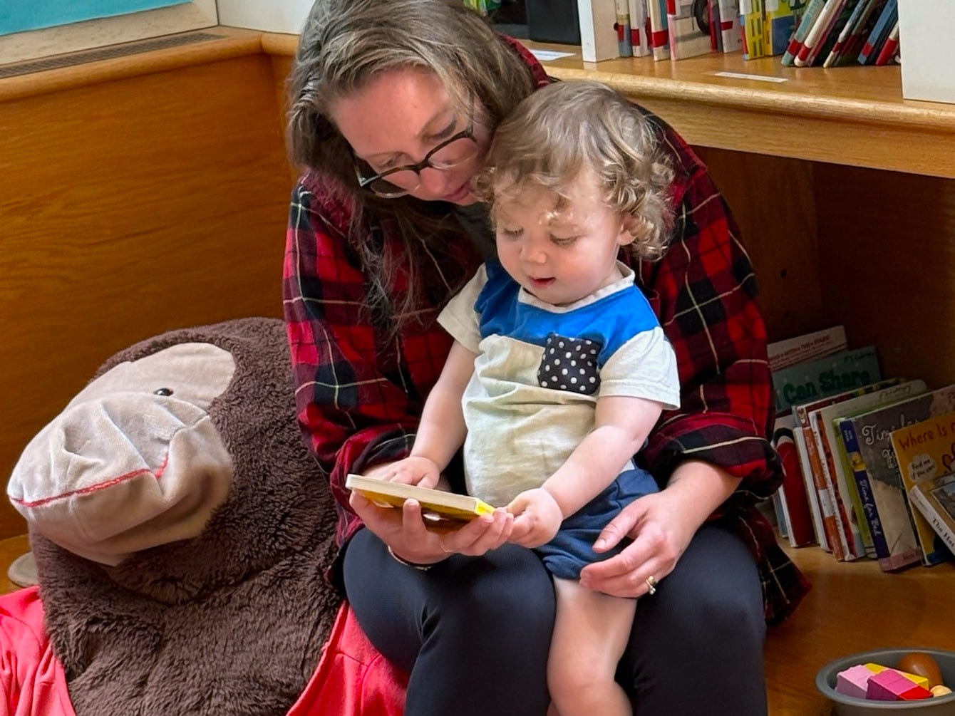A toddler sits on his mom's lap in a library, reading a board book.