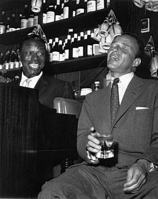 This may contain: two men in suits sitting at a bar with bottles on the shelves and one holding a drink This may contain: two men in suits sitting at a bar with bottles on the shelves and one holding a drink