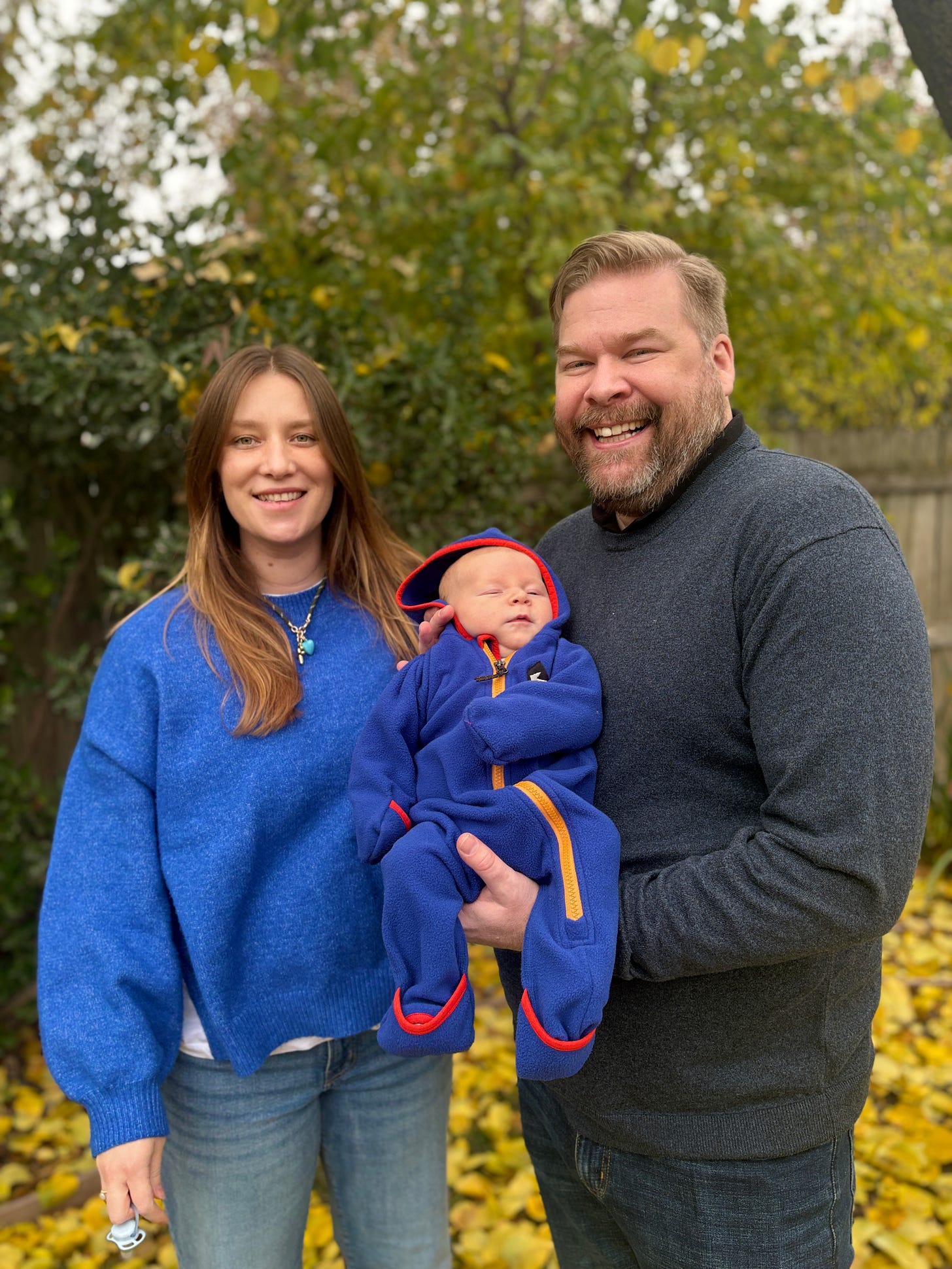 Cormac smiles alongside his wife and their infant son, posing together outdoors in a yard covered with autumn leaves.