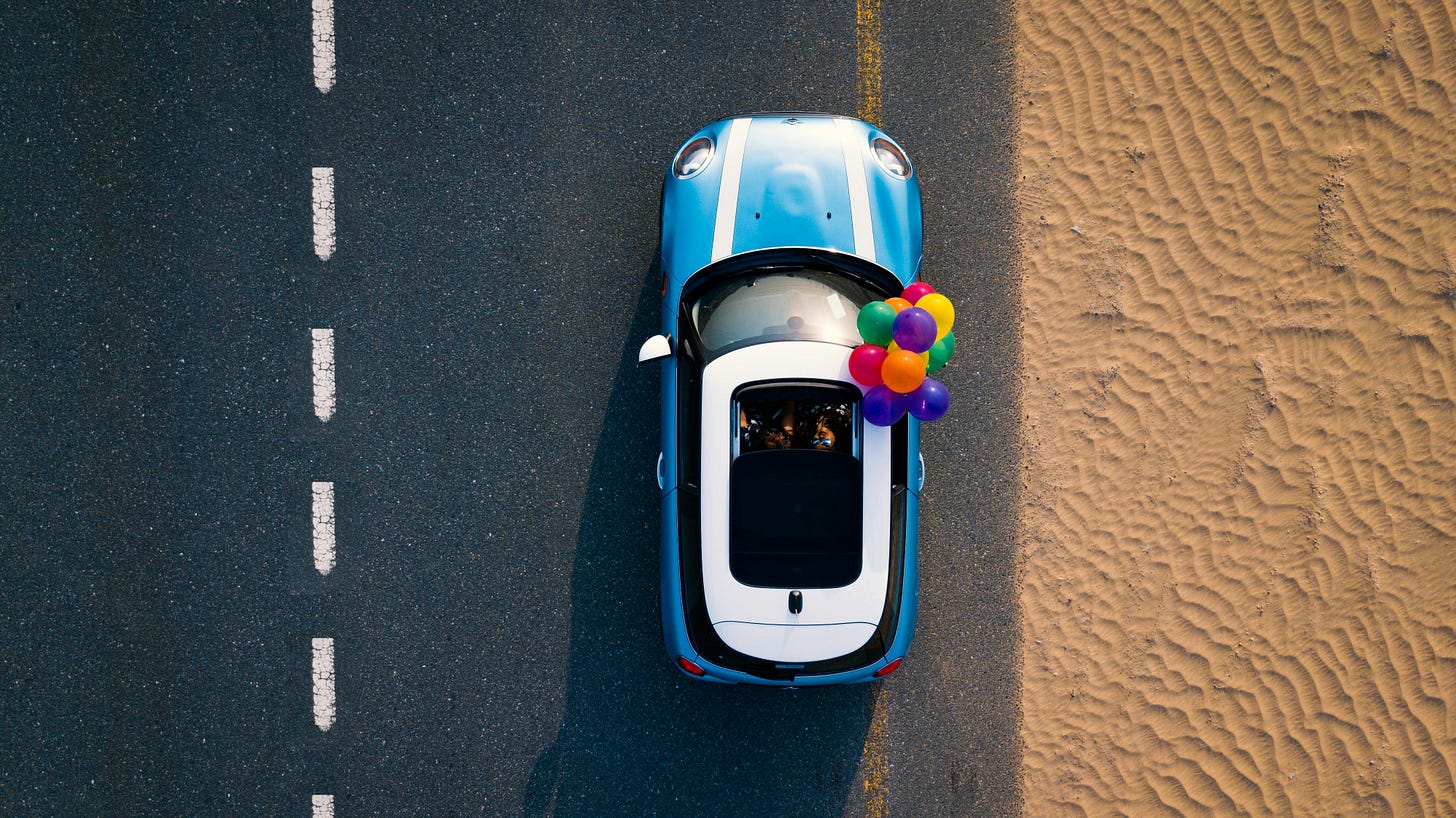 A foto mostra um carro azul visto de cima, estacionado ao lado de uma estrada. O carro tem um teto solar aberto e do lado direito dele há um grupo de balões coloridos amarrados. A estrada tem uma linha branca tracejada no centro e está ladeada por uma área arenosa, com pegadas visíveis na areia.