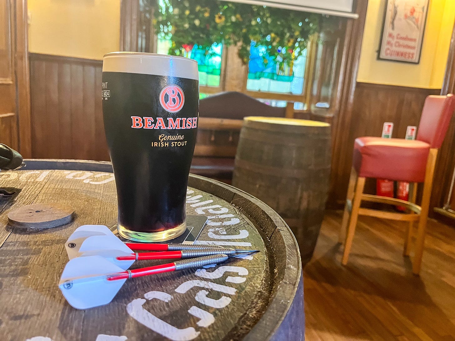 A full pint of stout in a Beamish branded glass, on top of a barrel next to three red and white darts.
