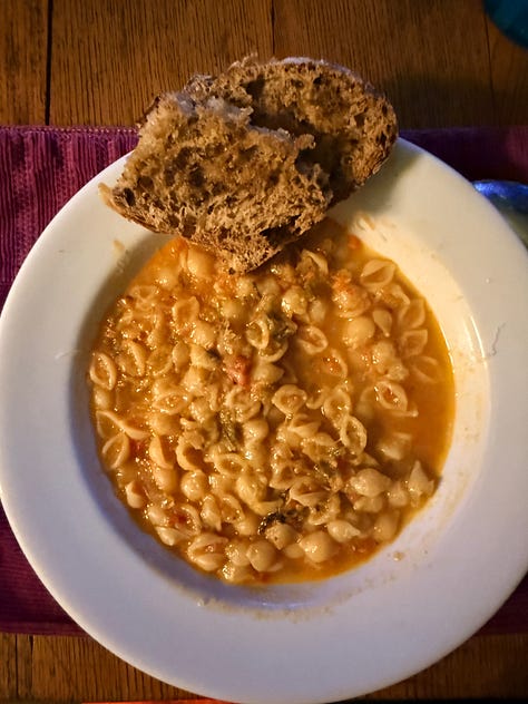 Image 1: A white bowl filled with small shell pasta in a tomato-based broth with vegetables, served on a wooden table. A torn piece of rustic brown bread rests on the rim of the bowl. Image 2: A blue-and-yellow box of De Cecco Small Shells No. 52 pasta, showing the pasta through a clear window on the front. The packaging highlights that it is slow‑dried for superior quality and made in Italy. Image 3: A large pot filled with a simmering mixture of diced vegetables—such as onions, celery, and red peppers—in a seasoned broth, likely the base for a soup or pasta dish.