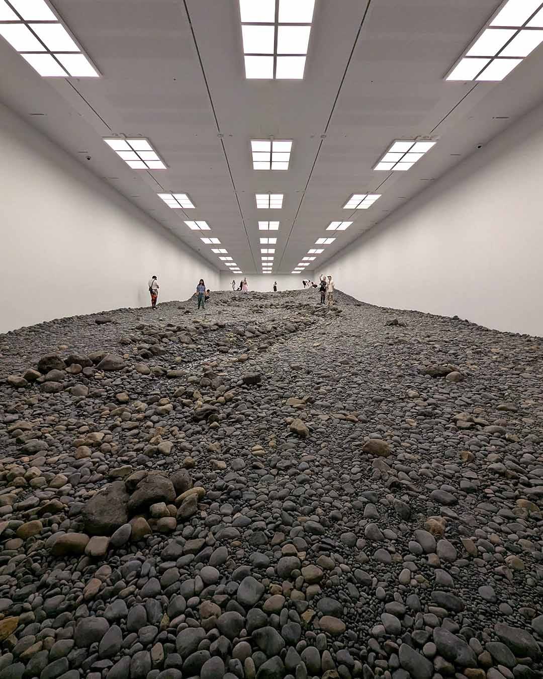 Large-scale installation of gray and brown volcanic rocks covering entire gallery floor creating steep hill or mountain formation, with visitors visible as small figures climbing or standing on rocky surface under bright grid ceiling lights in white gallery space