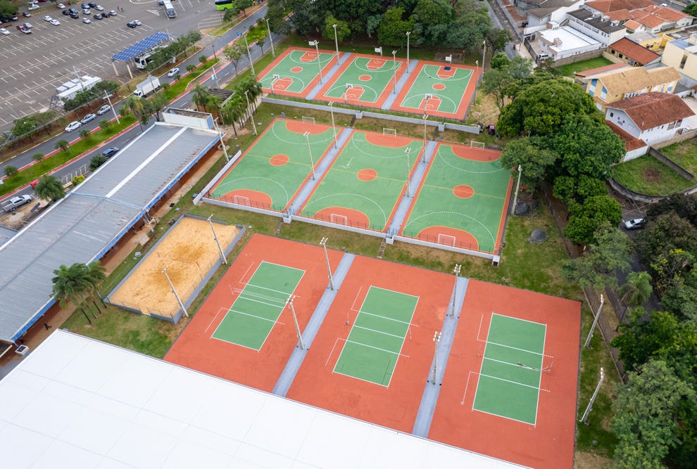 Vista aérea de um complexo esportivo contendo múltiplas quadras poliesportivas com superfície verde e laranja, três quadras de tênis com piso verde cercadas por área laranja, uma quadra de areia, além de estacionamento, áreas arborizadas e edificações adjacentes.