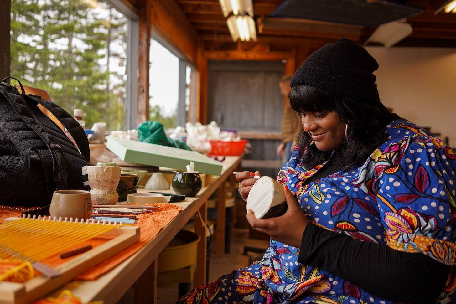 female-presenting person in colorful clothes works on a pottery piece amid creative clutter