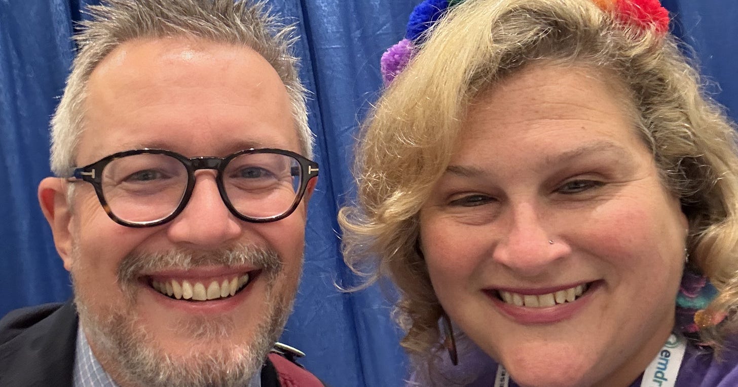 a white man with glasses, grey hair and a bowtie poses with a blonde woman wearing a rainbow bandana