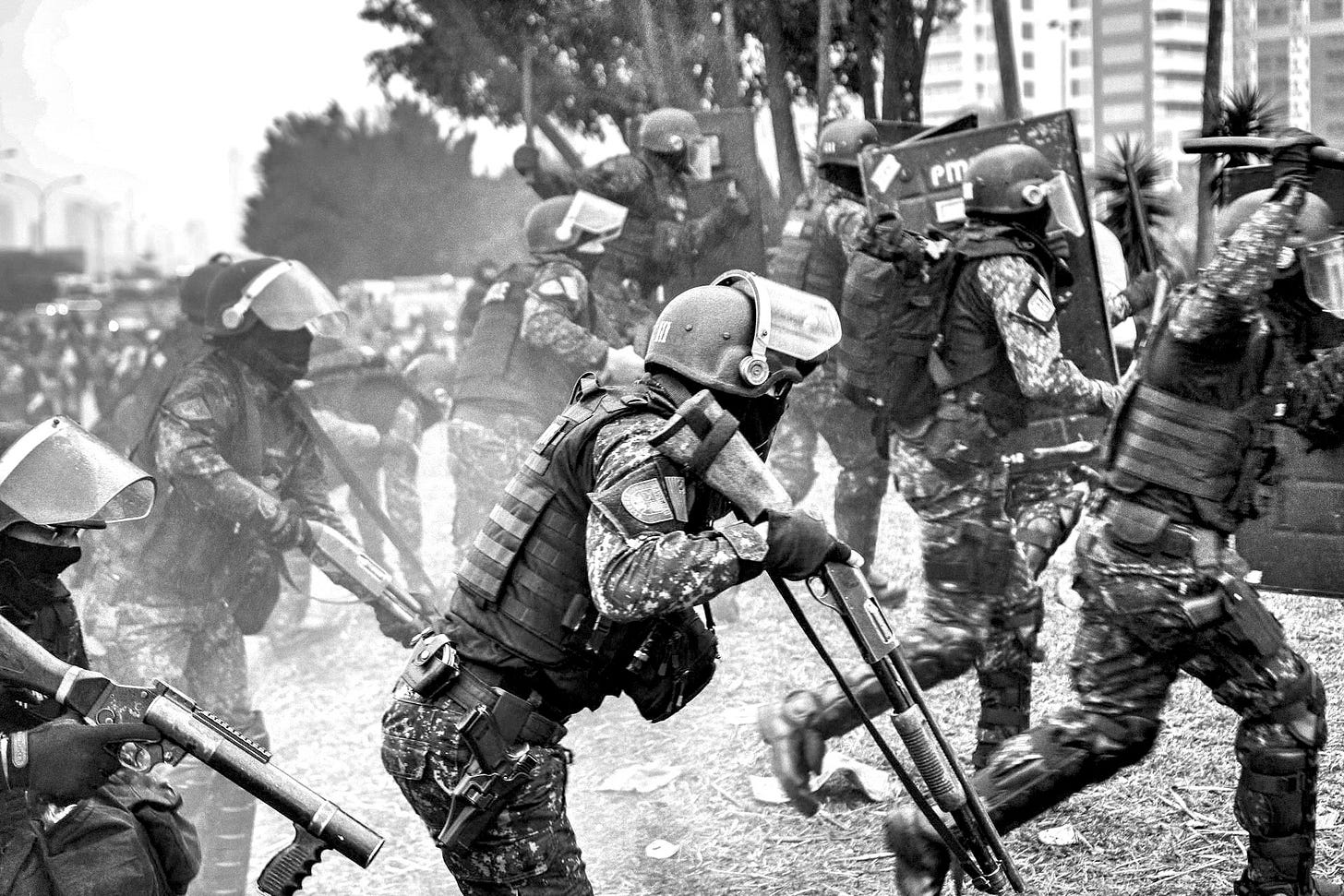 Riot police in full tactical gear advancing during what appears to be a protest. Officers wear black uniforms, helmets with face shields, and carry riot shields and weapons. The scene is smoky with urban buildings in the background. The image captures police in formation during a tense confrontation.