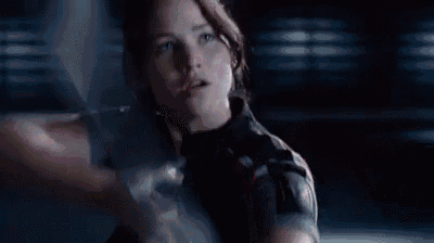 A woman intensely draws a bow in a dark setting, ready to release an arrow. She wears dark clothing, and the mood is focused and determined.