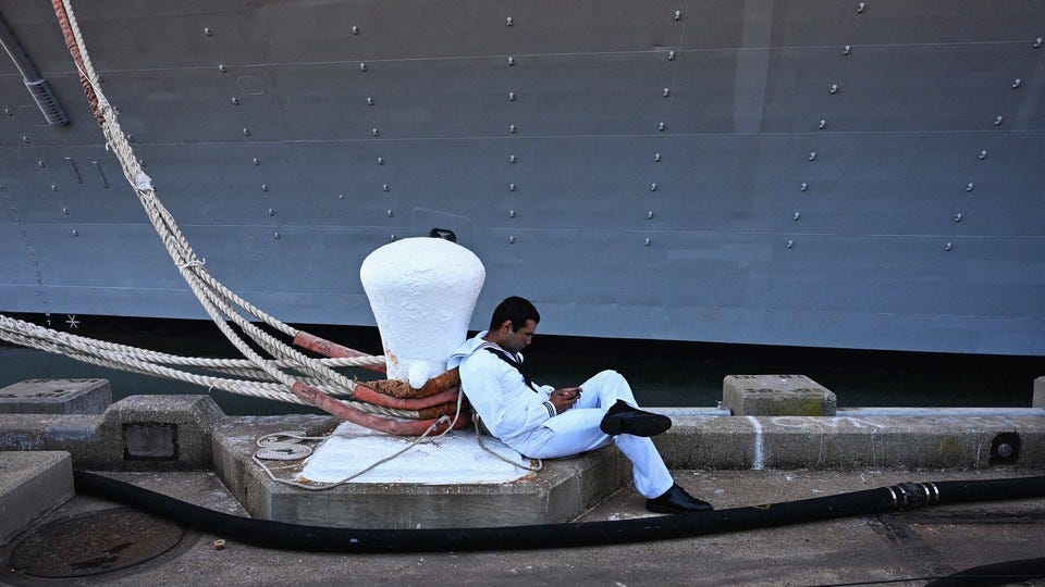 A sailor in a white uniform leans against a white bollard on a dock in front of a ship A sailor in a white uniform leans against a white bollard on a dock in front of a ship