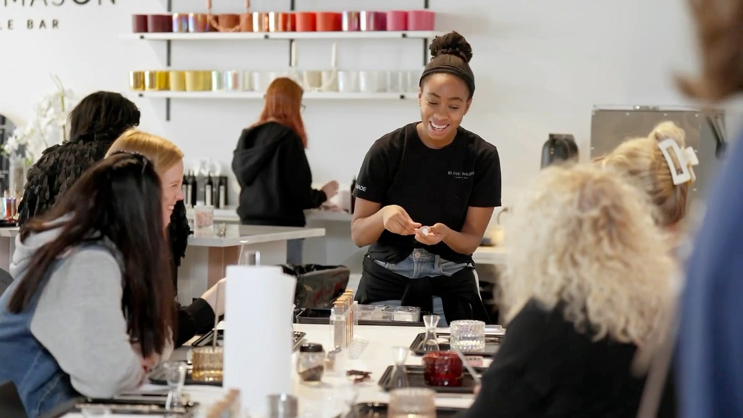A workshop session at Blank Mason Candle Bar showing a staff member demonstrating candle-making techniques to customers seated at a white countertop. Various candle-making supplies and test fragrance vials are arranged on the counter.