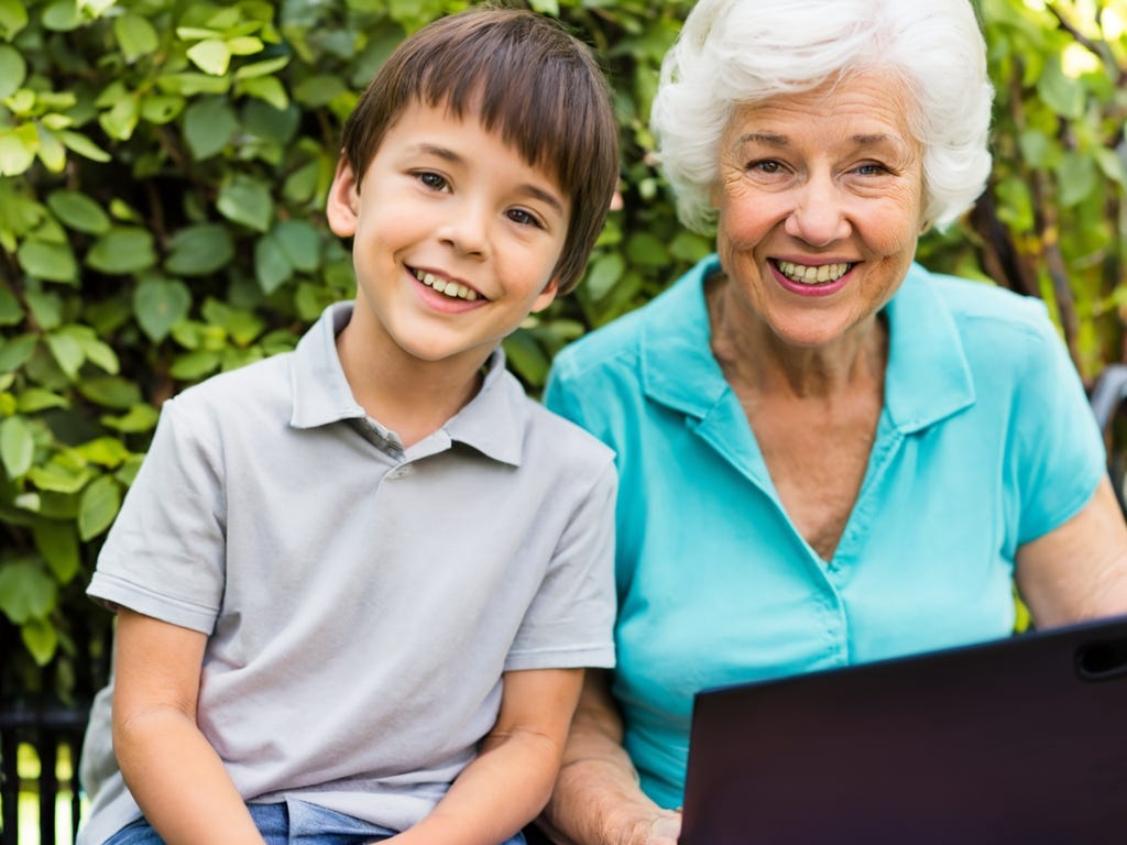 Both smiling, 82-year-old grandmother with white hair sits with 9-year-old grandson with garden in background. 