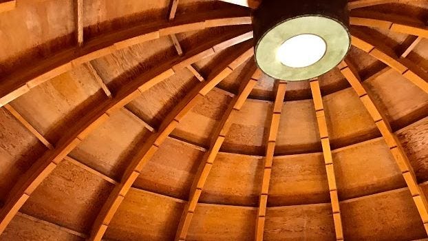 Image: A round skylight of a domed structure and the warm brown wooden panels of the ceiling.