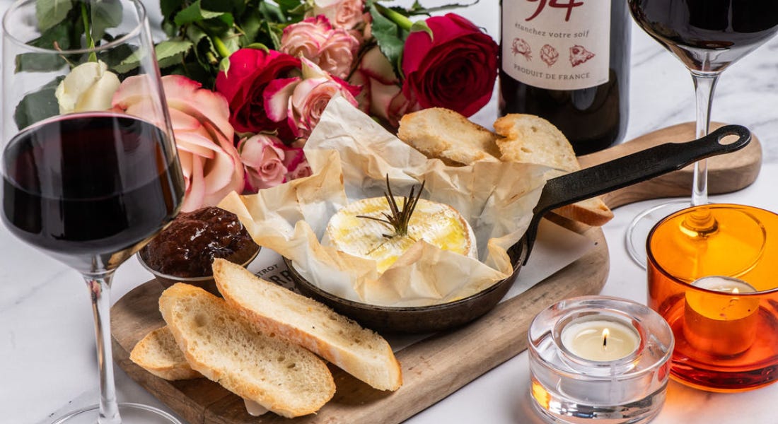 A grey marble table with a glass of red wine, a bunch of roses, a wooden board with camembert and bread on top of it, and candles on it