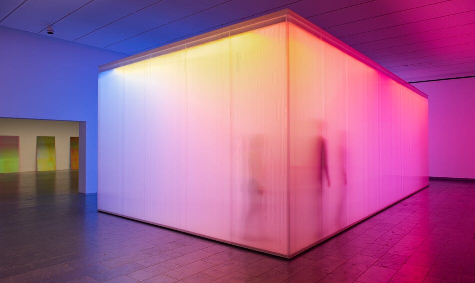 A cube in a gallery space the has semi translucent walls illuminated in pink, purple and yellow light, in which you can see the outline of people walking around inside.