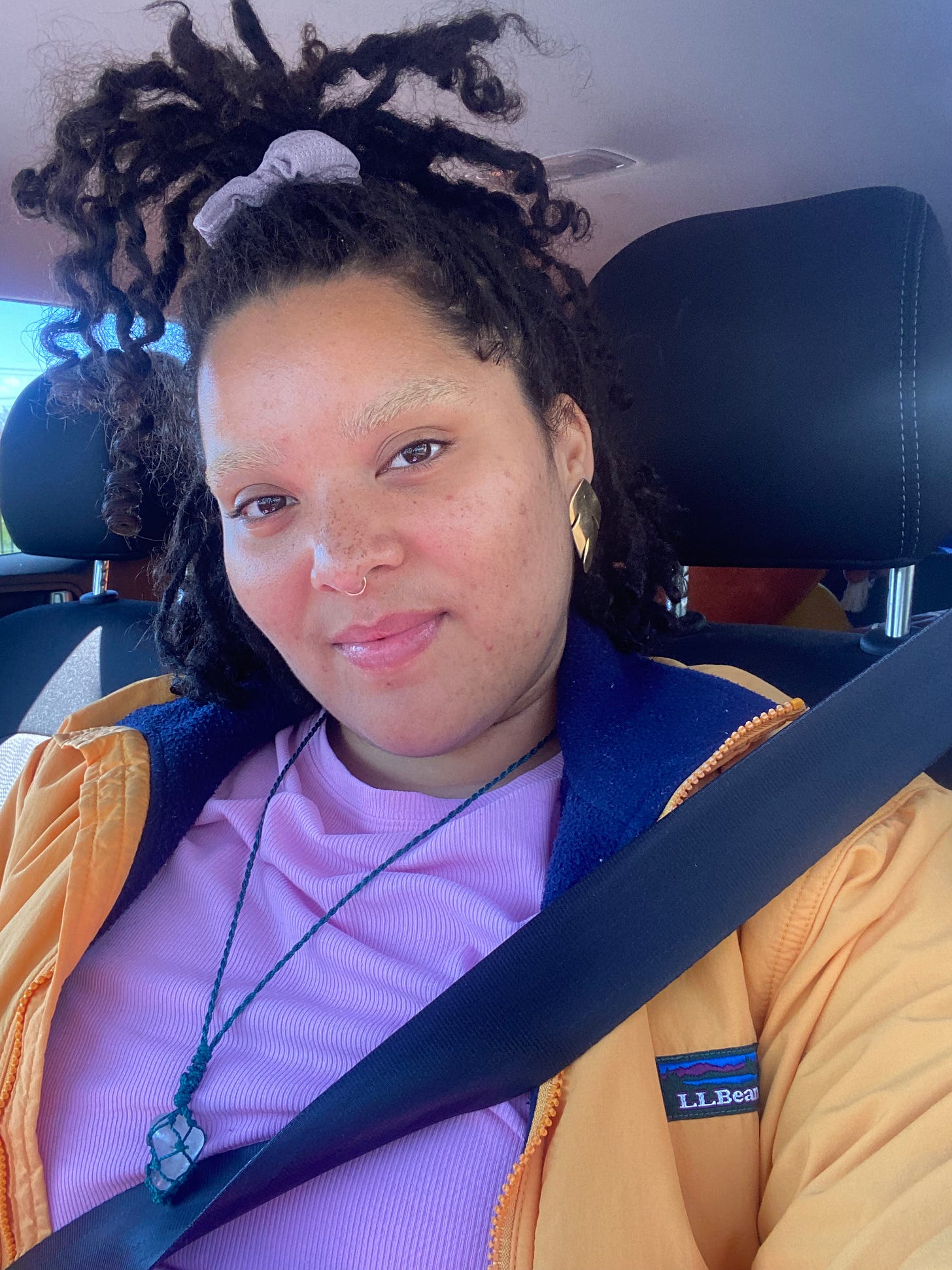 Kay wears hair half up half down while seated in the car wearing a pink top, yellow jacket and a smile.