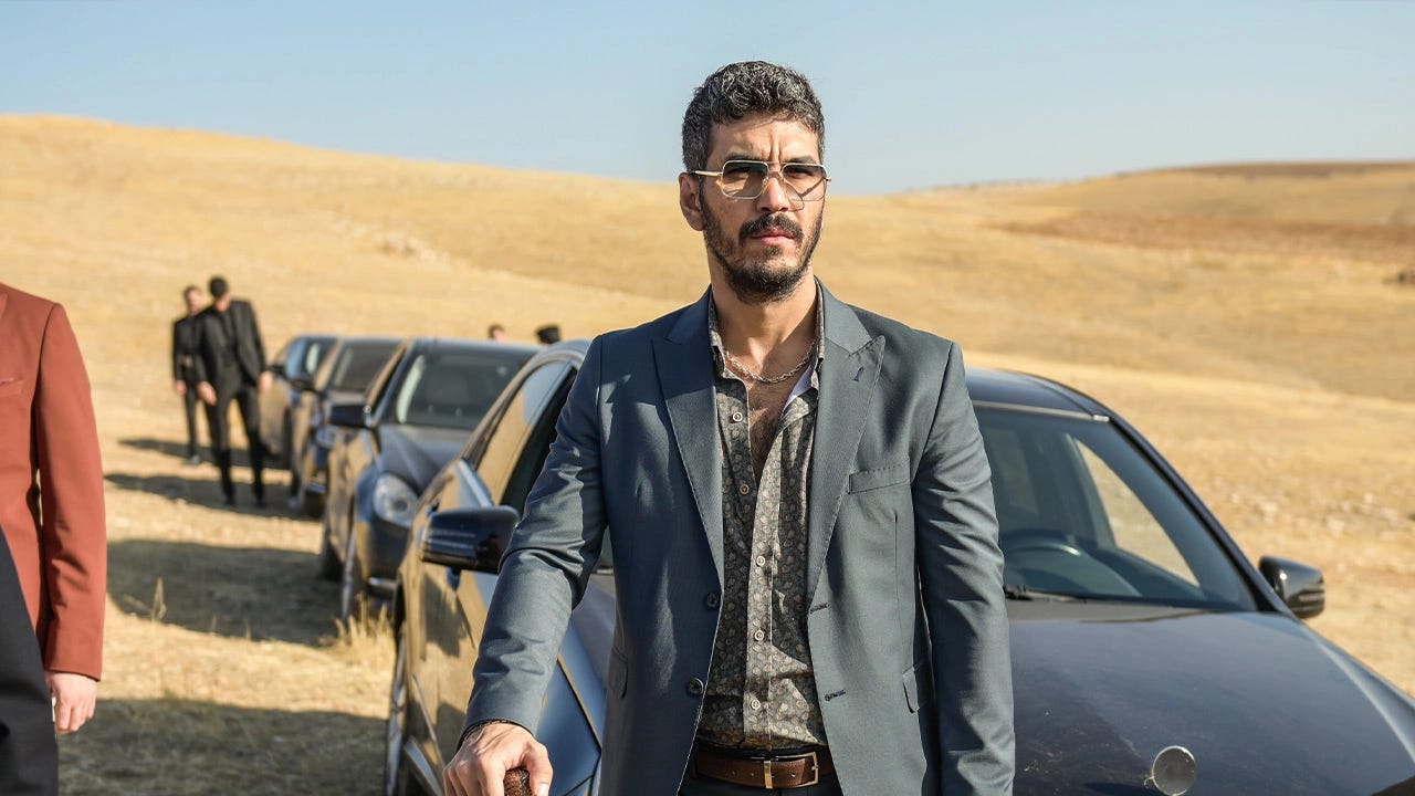 Halef: Köklerin Çağrısı dizisinde çöl yolunda siyah araç konvoyunun önünde duran takım elbiseli erkek karakter Halef: Köklerin Çağrısı dizisinde çöl yolunda siyah araç konvoyunun önünde duran takım elbiseli erkek karakter