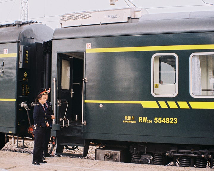 一张写实的铁路摄影。画面右侧是一节深绿色的 RW25G型软卧车厢，车身印有黄色编号 554823。两名身着蓝色制服的乘务员正站在车门旁的站台上值守。车顶的设备带有 Guangzhou (广州) 的字样。照片色调柔和，捕捉了普速列车运行中的一个静谧瞬间。A realistic railway photograph featuring a dark green RW25G (Type 25G Soft Sleeper) carriage with the yellow number 554823 on its side. Two train attendants in blue uniforms are on duty on the platform next to the carriage door. The equipment on the roof bears the name Guangzhou (广州). The photo has a soft tone, capturing a quiet moment of a conventional train in service.