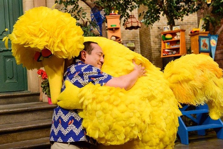 Big Bird the muppet, a large yellow bird, hugs a man in a blue shirt with zig zag stripes in front of a stoop on Sesame Street