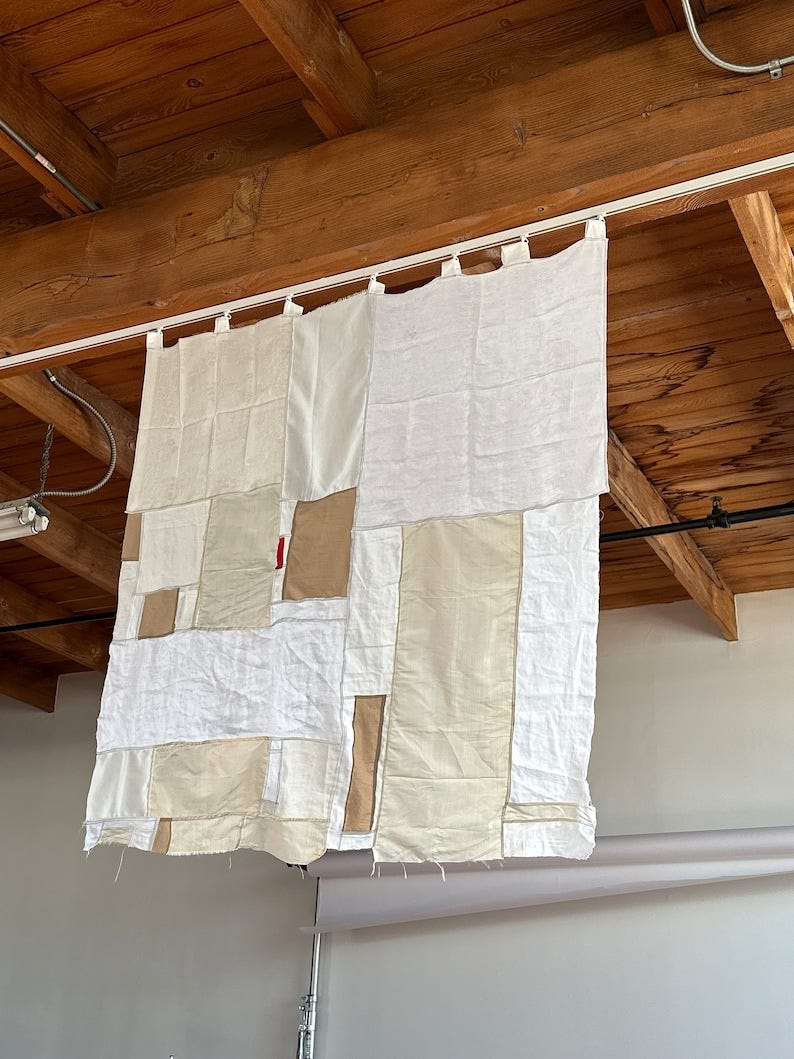 May include: A white and beige patchwork curtain hanging from a wooden ceiling beam. The curtain is made of different sized squares of fabric sewn together.