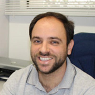 Homem de barba sorridente usando camisa cinza sentado em frente a mesa com computador, segurando uma caneta Homem de barba sorridente usando camisa cinza sentado em frente a mesa com computador, segurando uma caneta