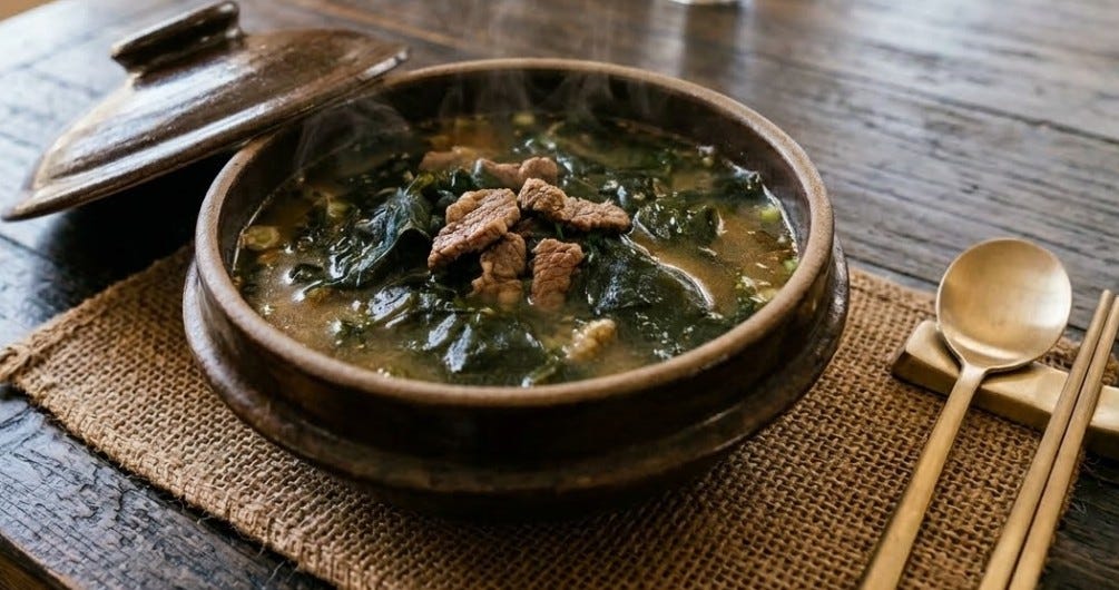 A close up photograph of a steaming ceramic bowl filled with dark green Korean seaweed soup (miyeok-guk) topped with several pieces of cooked beef. The bowl is placed on a burlap mat over a rustic dark wooden table. Traditional Korean brass utensils, including a spoon and chopsticks, are neatly arranged on a rest to the right of the bowl, and the ceramic lid of the bowl rests partially open in the background.