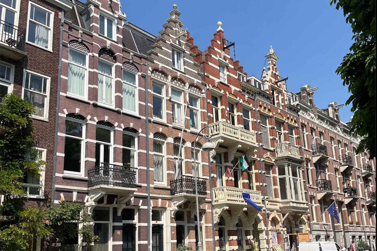 A row of ornate, historic townhouses in Amsterdam with stepped gables and decorative facades, photographed on a sunny day with clear blue skies. Trees partially frame the right edge of the image.