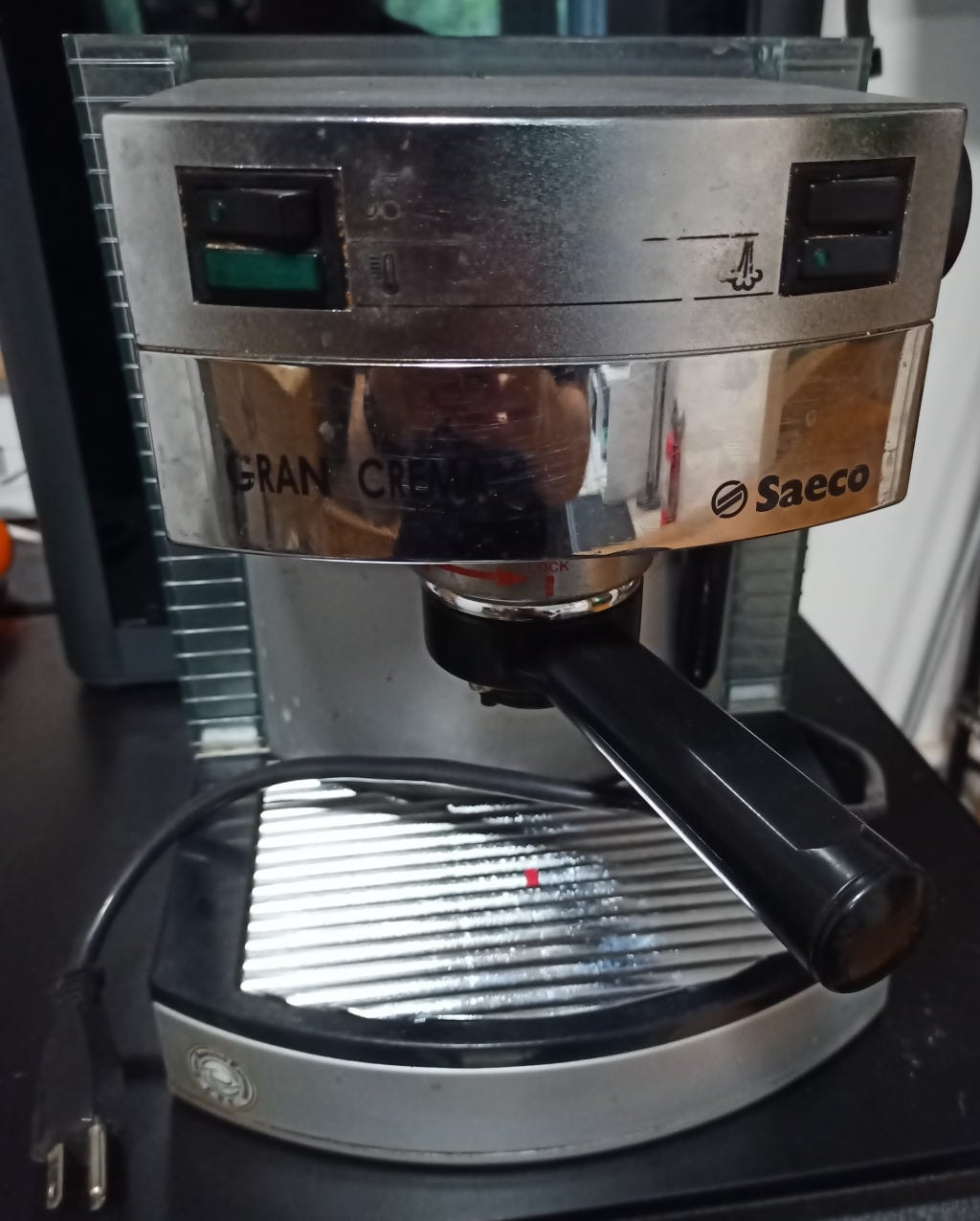 A small espresso/cappuccino machine. At the top left there is a black on/off switch; the markings to indicate the purpose are barely visible. Below that is a green light of the same proportions. The icon beside the light indicates that this means the machine is at operating temperature. on the right, there are two switches. The icon for the top switch (to turn the pump on) is completely obliterated. the switch below has a "steam" icon, and a green lamp is embedded into the switch. At the rear of the unit, the water tank wraps around both sides, the tank has many horizontal lines that make it easy to judge the water level.