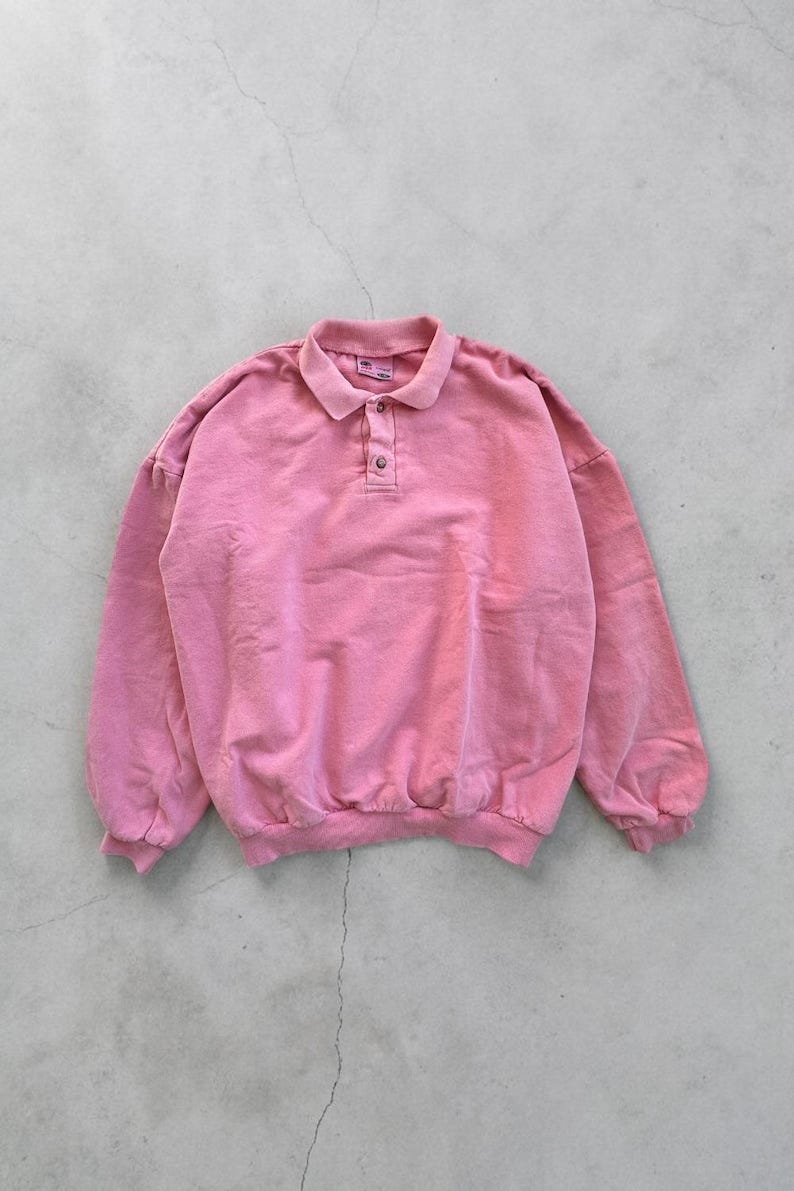 May include: A pink polo-style sweatshirt with a collared neckline and three buttons. The long-sleeved top has a relaxed fit and a ribbed hem and cuffs. The fabric appears to be a soft, faded material.