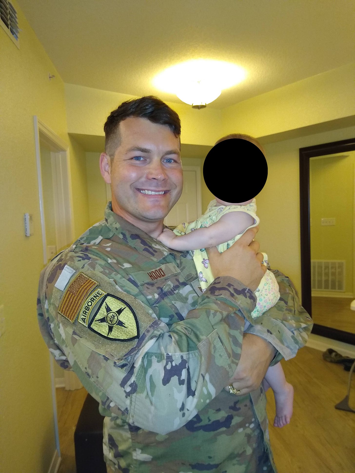SSgt Edward Hood smiling in his camouflage uniform holding his daughter indoors. SSgt Edward Hood smiling in his camouflage uniform holding his daughter indoors.
