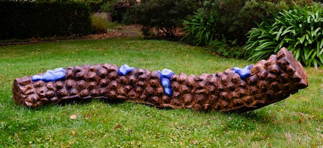 Long horizontal redwood sculpture placed outdoors on grass, partially charred and painted, with a textured surface of repeating rounded protrusions and several blue-painted sections. Long horizontal redwood sculpture placed outdoors on grass, partially charred and painted, with a textured surface of repeating rounded protrusions and several blue-painted sections.