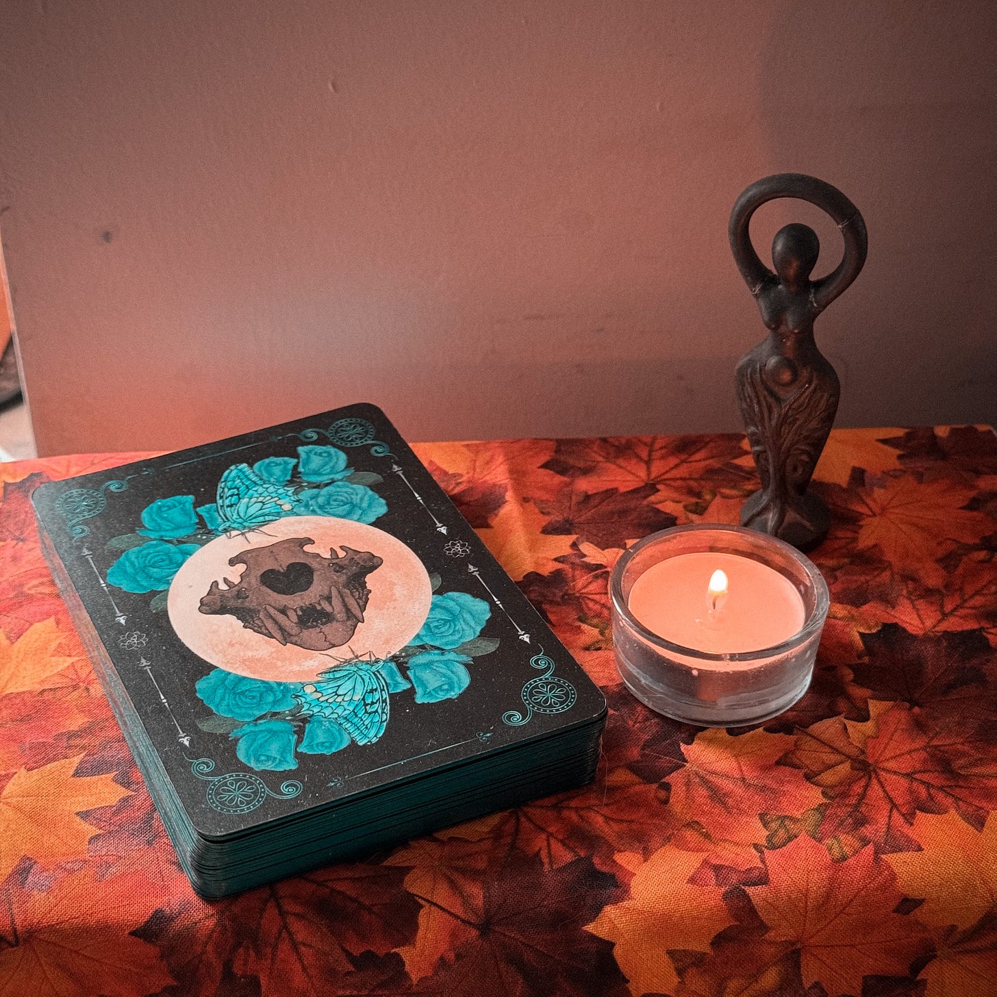 An oracle deck, tea light, and goddess statue stand on an altar cloth. The tea light is lit, casting light onto the goddess. The oracle deck is black and shows a card back featuring blue roses and butterflies around the skull of a carnivore. The altar cloth is richly patterned in red, gold, and brown autumn leaves.