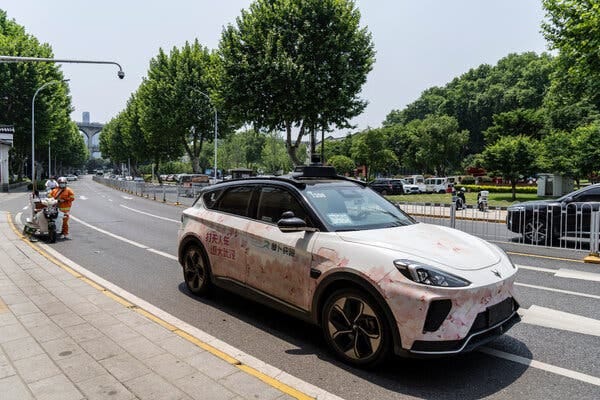 A white car with rose colored flowers painted on it on a city street. Behind it a man wearing an orange work outfit and helmet stands next to a motorbike