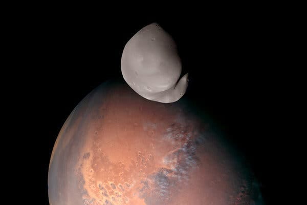 The small moon Deimos, which is gray and oblong and partially cast in shadow, appears to hover over the large red orb of Mars against the blackness of space. The small moon Deimos, which is gray and oblong and partially cast in shadow, appears to hover over the large red orb of Mars against the blackness of space.