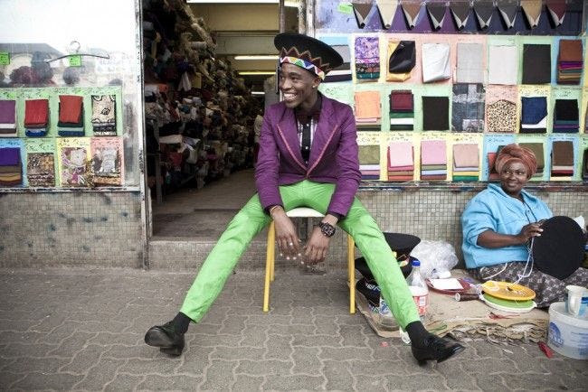 This may contain: two people sitting on chairs in front of a store with lots of colorful fabrics and hats