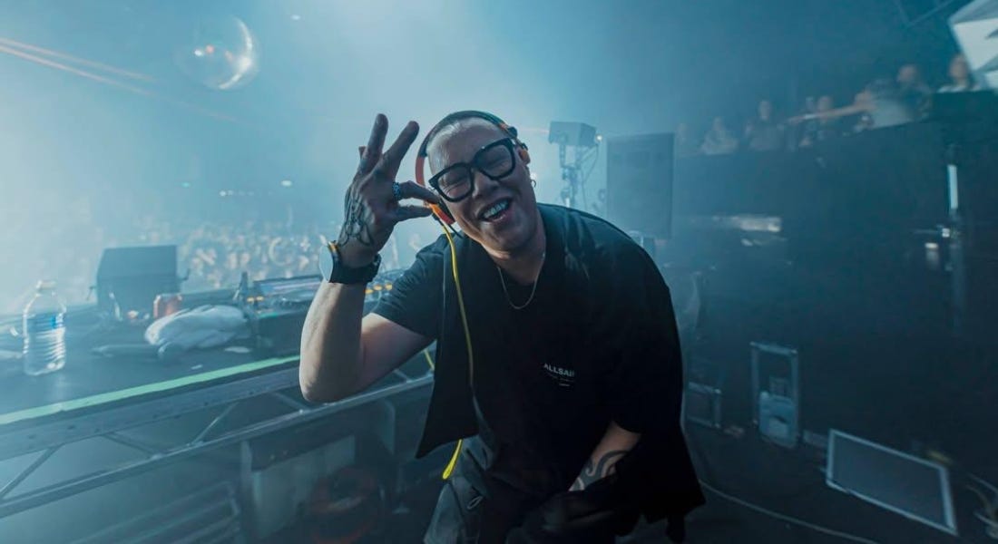 Gok Wan wearing glasses and a black t-shirt smiling into the camera, with DJ decks and an audience behind him