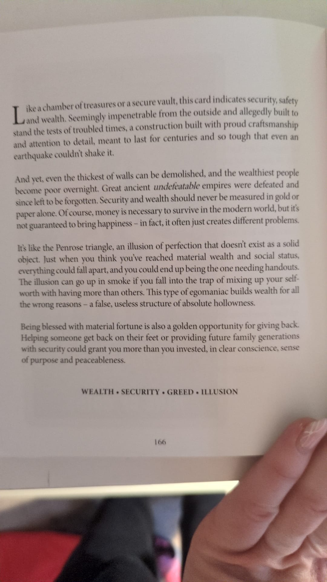 A photograph of an open tarot guidebook page discussing the Ten of Coins. The text reflects on wealth, security, illusion, and generosity, comparing material stability to a fortified structure and the Penrose triangle. At the bottom of the page, the keywords “WEALTH · SECURITY · GREED · ILLUSION” are printed above the page number 166.