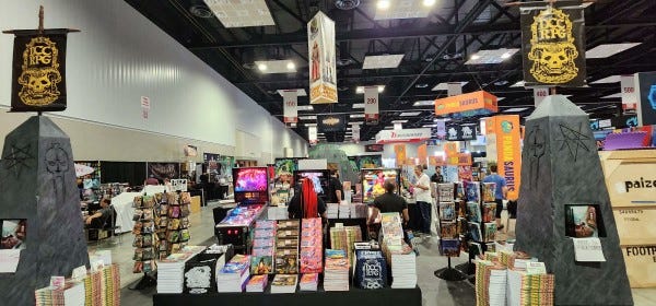 A wide shot of Goodman Games' booth at Gen Con 2023. There are obelisks, a ziggurat, and pinball tables among the stacks of books.