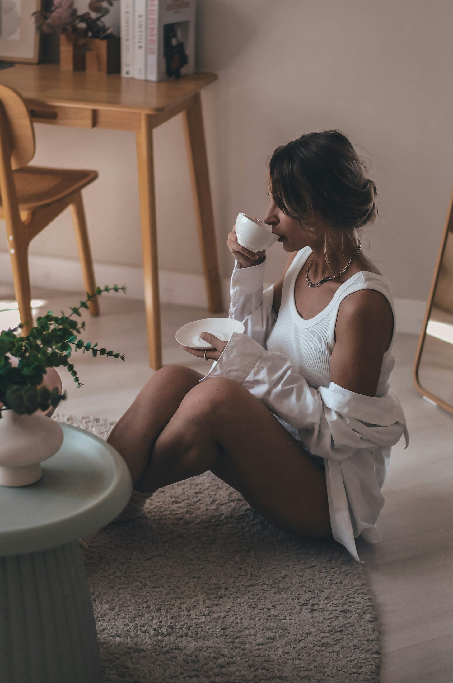 A woman sits on a round rug, drinking from a teacup and saucer, wearing a white shirt and tank top, with a table, chair, and plant in the background. A woman sits on a round rug, drinking from a teacup and saucer, wearing a white shirt and tank top, with a table, chair, and plant in the background.