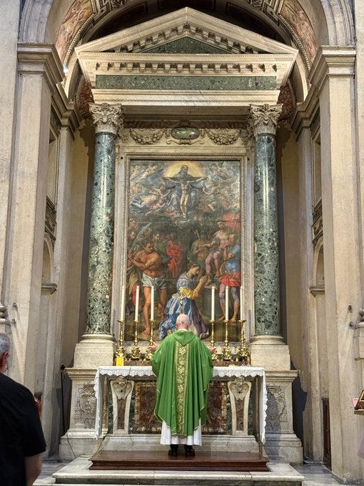 A priest in green vestments stands at an altar in a grand basilica. The altar features ornate columns, a large painting of a crucifixion scene with figures, and candelabras with lit candles. Marble and decorative stonework frame the scene, with an arched architectural structure above.