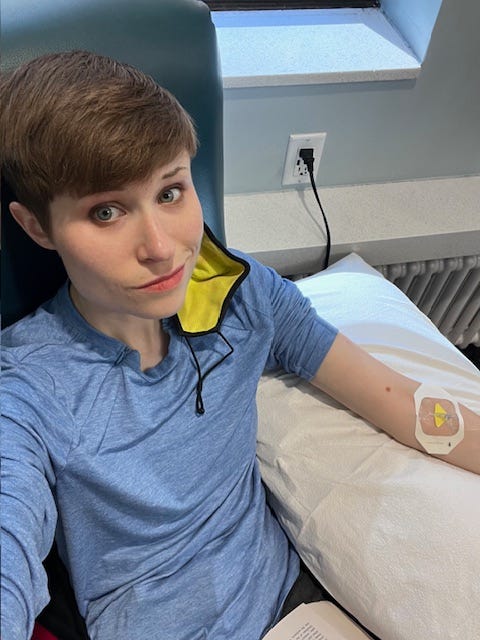 Selfie of writer in infusion chair, blue mask dangling from one ear. There's an IV in their propped left arm and an open book on their lap.