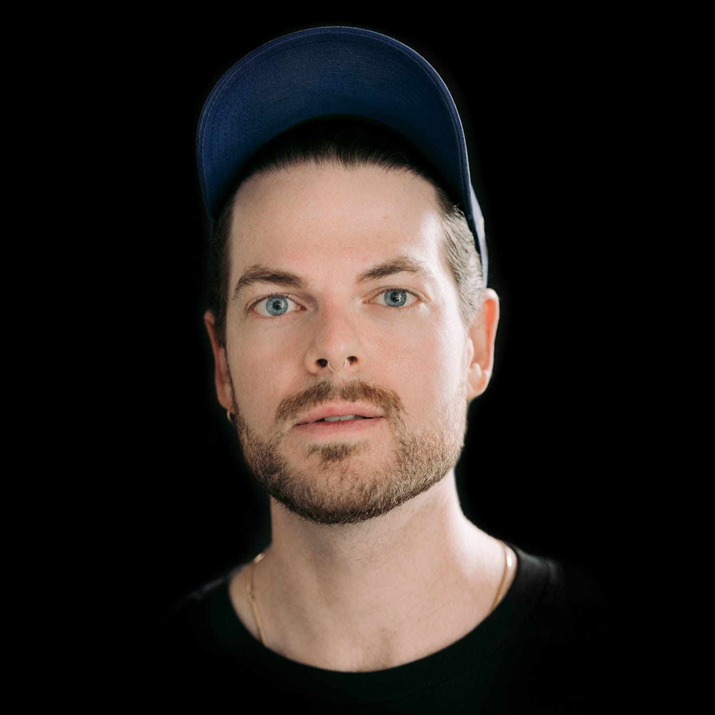 A twentysomething white man with brown facial hair and light blue eyes looks into the camera. He wears a navy cap, an earring in his right ear and a thin gold necklace.