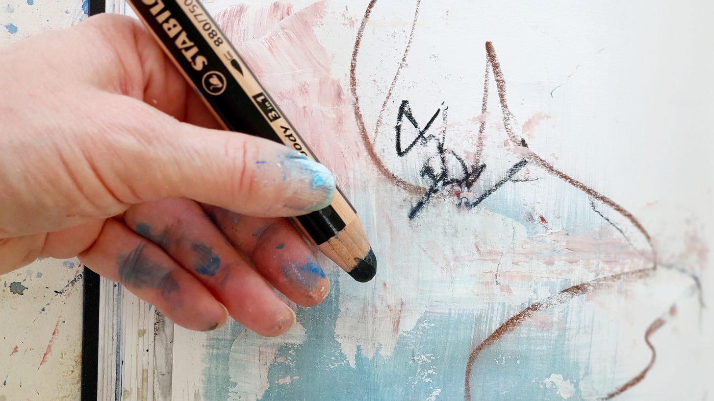 the artist Susan Mills holding a thick Woody pencil and drawing over paint in a sketchbook. Hand is painted too.
