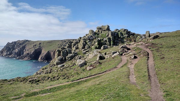 A photograph of a rugged coastal landscape of grass, rocks and a path, with a glimpse of sea to the left, under a nice blue sky with a little cloud to the left.