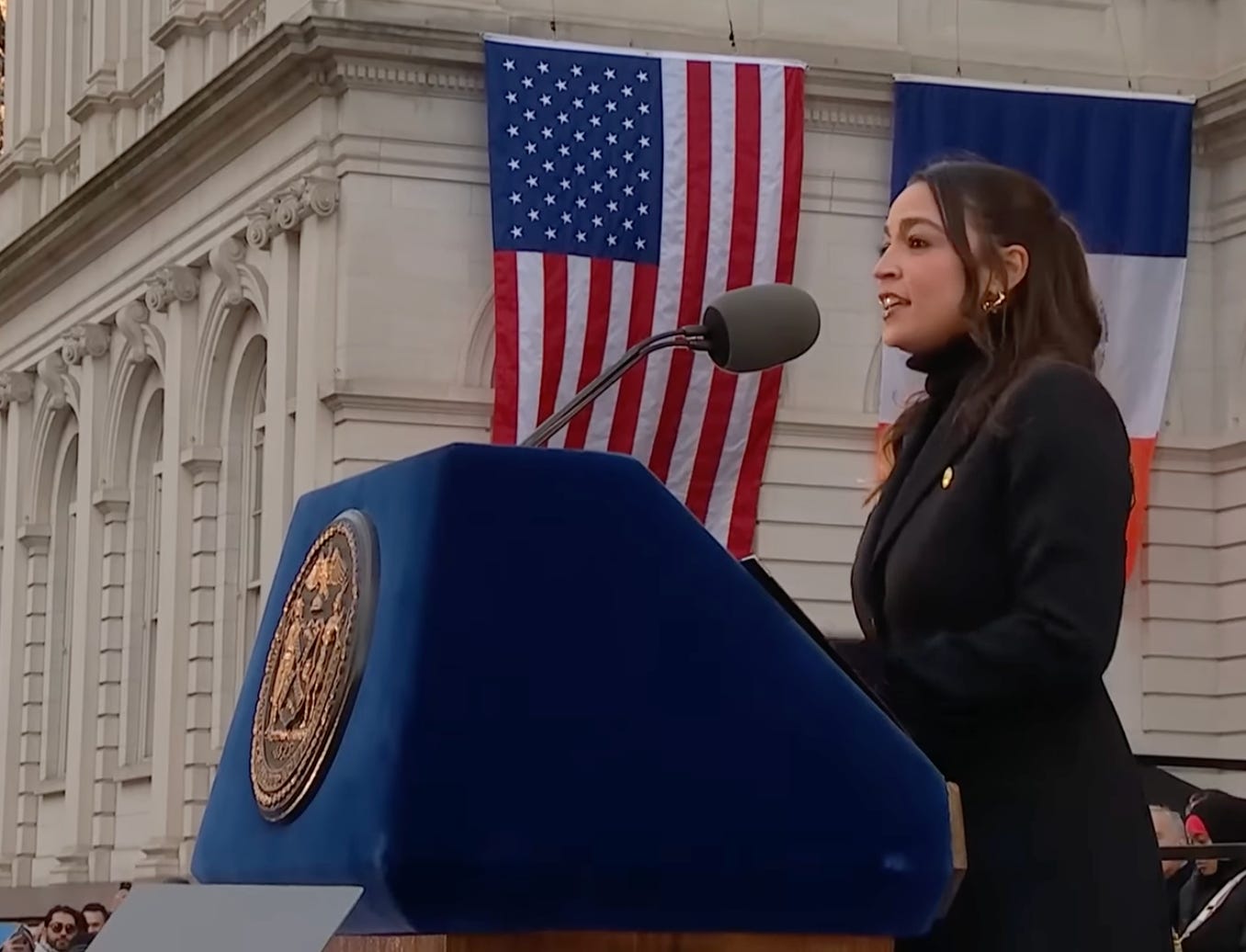 AOC in profile at podium; classical building draped with flag behind her