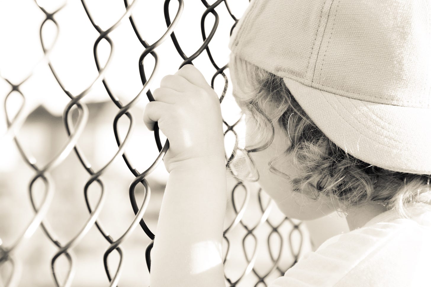 a young child, wearing a backwards baseball cap, grasps onto a chain-link fence, staring out onto what is on the other side of the fence (which is out of focus)