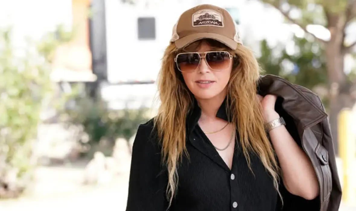 blond woman wearing a black shirt, a brown baseball cap and big eyeglasses holds a leather jacker