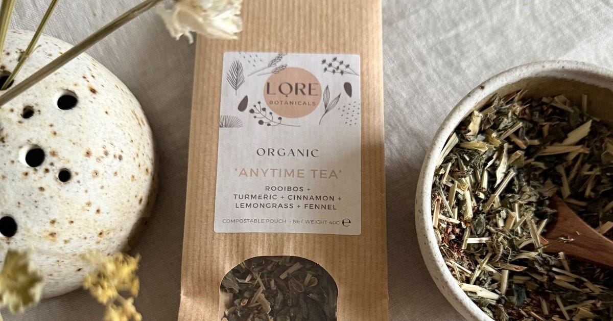 Lore Botanicals Anytime Tea - photographed in its packaging which is brown kraft paper with an illustrated label and a small window where the herbs are visible. Next to this is a bowl of herbs and some flowers