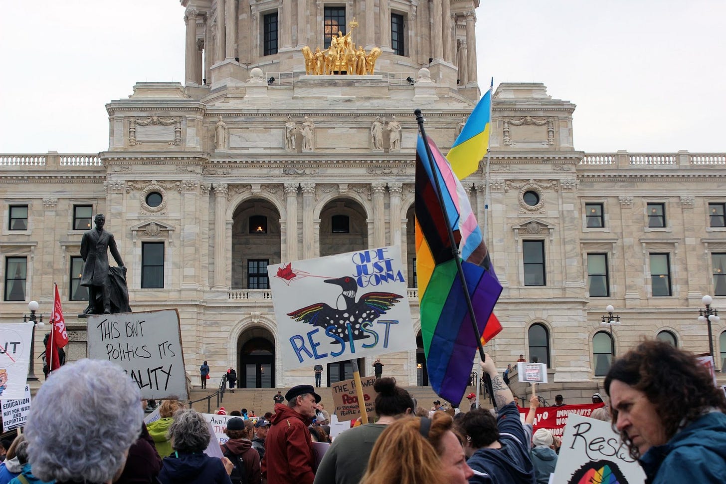 a crowd gathers in front of the state capitol, one sign depicts a loon with lasers shooting out of its eyes and reads "RESIST" a crowd gathers in front of the state capitol, one sign depicts a loon with lasers shooting out of its eyes and reads "RESIST"