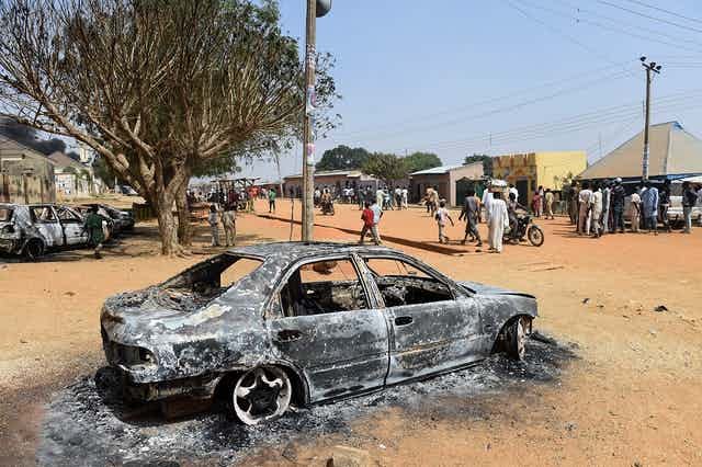 Burnt out car on an unpaved road, with buildings and people in the background Burnt out car on an unpaved road, with buildings and people in the background
