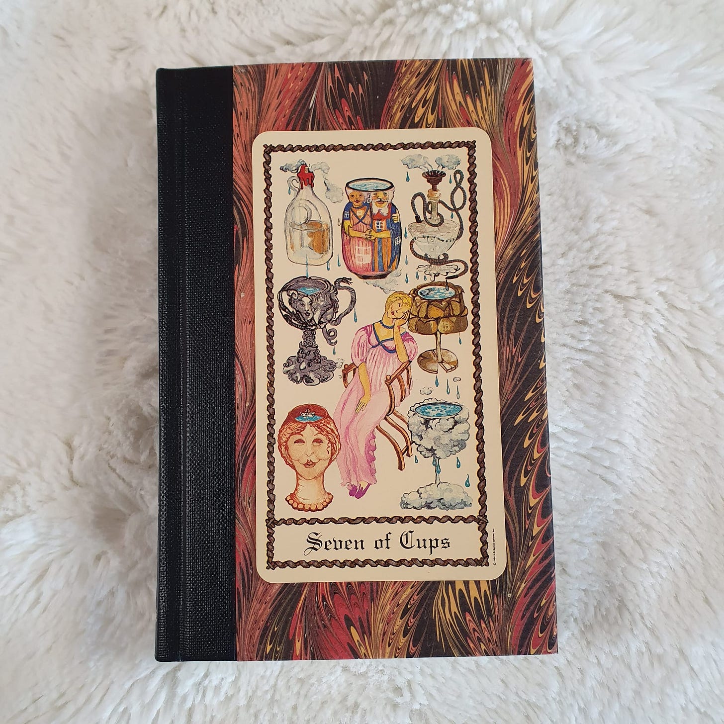 Seven of Cups card from the Scapini Tarot displayed on a marbled book cover Seven of Cups card from the Scapini Tarot displayed on a marbled book cover
